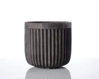 En mørkegrå, cylindrisk beholder med mat finish og lodrette riller, der løber fra bunden til kanten. Kanten er en smule tykkere og afrundet, hvilket giver et robust udtryk. Overfladen har et stenlignende udseende, hvilket antyder, at den er lavet af beton eller et lignende materiale.