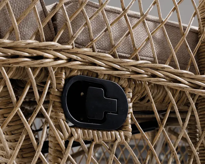 Nærbillede af en ramme i flettet rattan med en beige hynde ovenpå. Rammen har et indbygget sort plastikhåndtag eller greb, sandsynligvis til at justere positionen eller liggefunktionen. Rattanen er tæt flettet i et krydsmønster, og hynden har et tekstureret stofbetræk.