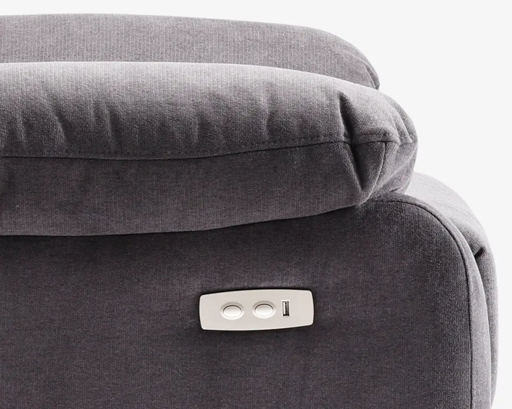 Nærbillede af en mørkegrå polstret overflade med en blød hovedstøtte og et integreret betjeningspanel. Betjeningspanelet har to ovale knapper og en USB-port, som muliggør elektronisk justering og opladning af enheder. Stoffet ser blødt ud, og syningerne er pæne, hvilket giver et komfortabelt og moderne udtryk.
