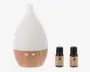 En dråbeformet enhed med mat hvid top og lys træstruktur i bunden, der har to hvide knapper. Ved siden af står to små brune flasker med sorte låg og beige etiketter, som angiver forskellige duftolier.