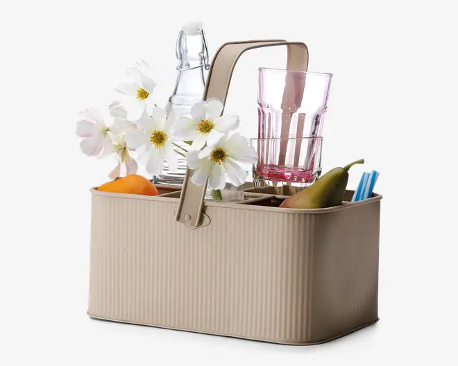 Rektangulær beige opbevaringskurv med rillet overflade og solidt centralt håndtag. Kurven er opdelt i rum og indeholder forskellige genstande såsom en glasflaske, et lyserødt drikkeglas, hvide kunstige blomster, en appelsin, en pære og noget bestik. Designet er praktisk til organisering og transport af køkken- eller picnicudstyr.