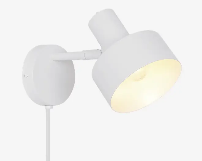 En moderne vægmonteret lampe med mat hvid finish. Lampen har en cylindrisk skærm, der kan vinkles for justerbar lysretning. Den har en rund baseplade til sikker montering på væggen og en synlig ledning, der går ud fra bunden. Designet er minimalistisk og passer godt til moderne indretning.