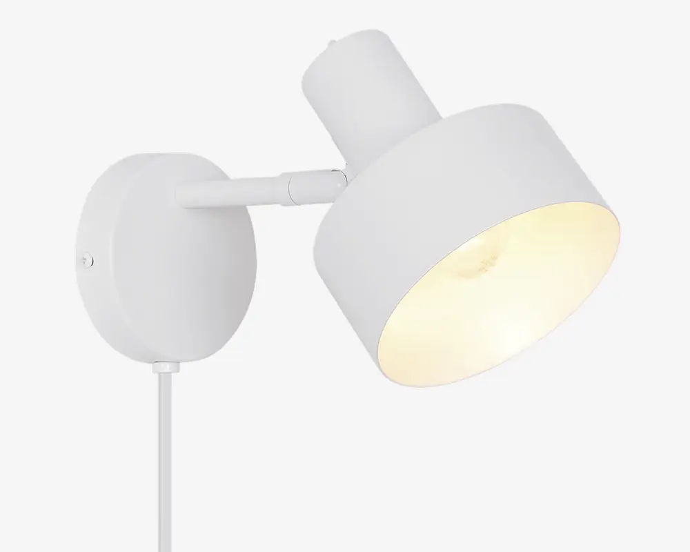 En moderne vægmonteret lampe med mat hvid finish. Lampen har en cylindrisk skærm, der kan vinkles for justerbar lysretning. Den har en rund baseplade til sikker montering på væggen og en synlig ledning, der går ud fra bunden. Designet er minimalistisk og passer godt til moderne indretning.