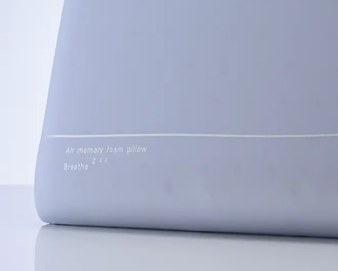 Nærbillede af en lysegrå pude med en glat, sømløs overflade og en diskret hvid linje, der løber vandret nær bunden. Puden har hvid tekst trykt med ordene "Air memory foam pillow" og "Breathe Zzz," hvilket indikerer, at den er lavet af air memory-skum for øget åndbarhed og komfort. Designet er minimalistisk og moderne.