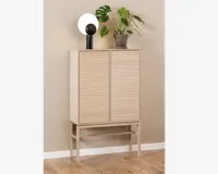 Høj skænk i lyst træ med to lodrette rillede låger, stående på slanke ben med en vandret støttebjælke. Skænken har et minimalistisk, moderne design og en glat, mat overflade. Ovenpå står en rund, hvid lampe med sort fod samt en grøn potteplante med store blade.