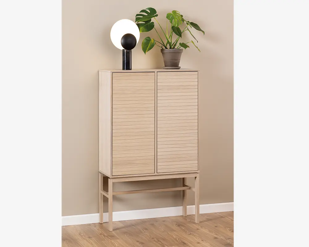 Høj skænk i lyst træ med to lodrette rillede låger, stående på slanke ben med en vandret støttebjælke. Skænken har et minimalistisk, moderne design og en glat, mat overflade. Ovenpå står en rund, hvid lampe med sort fod samt en grøn potteplante med store blade.
