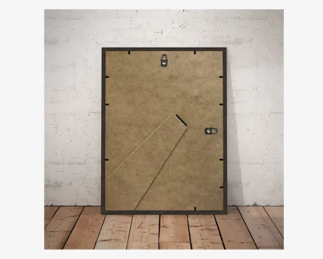 Billedet viser bagsiden af en rektangulær ramme med brun MDF-bagside. Der er en udklappelig støtte, som gør det muligt at stille rammen på et bord, placeret diagonalt fra nederste venstre hjørne. Rammen har to metalophæng: ét i toppen til lodret ophængning og ét på højre side til vandret ophængning. Rammen har en tynd, mørk kant.