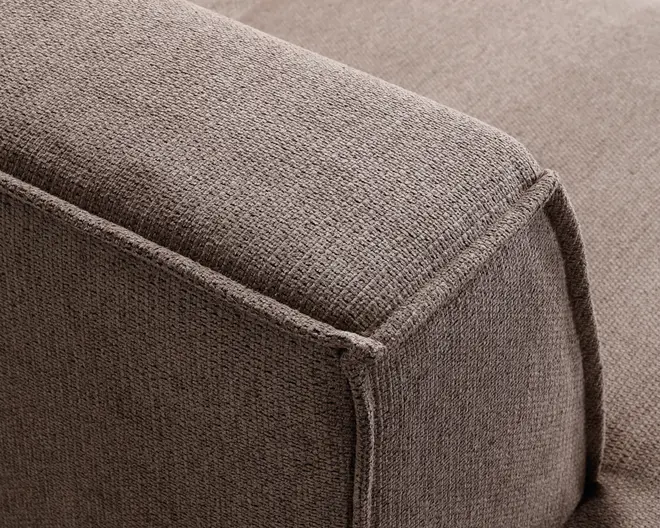 Nærbillede af en sofa-armlæn med tekstureret, tæt vævet brunt stofbetræk. Syningerne er synlige langs kanterne, hvilket giver et rent og struktureret udseende. Stoffet virker slidstærkt, og polstringen ser fast ud, hvilket giver både komfort og støtte.