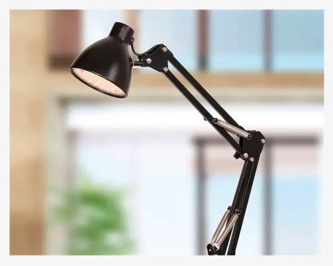 Sort justerbar skrivebordslampe med kuppelformet metalskærm, fjederbelastede led for fleksibel positionering og mat finish. Lampens hoved er vinklet nedad, og armen har synlige metalfjedre og samlinger for stabilitet og nem justering.