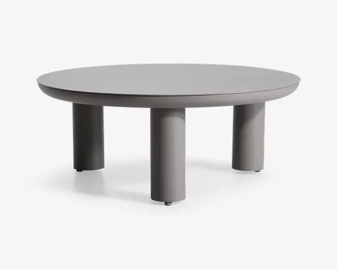 Rundt bord med en glat, mat grå overflade og fire tykke, cylindriske ben. Designet er minimalistisk og moderne med rene linjer og et solid, robust udtryk. Benene er jævnt fordelt og placeret tæt på kanten, hvilket giver stabilitet og et nutidigt look.