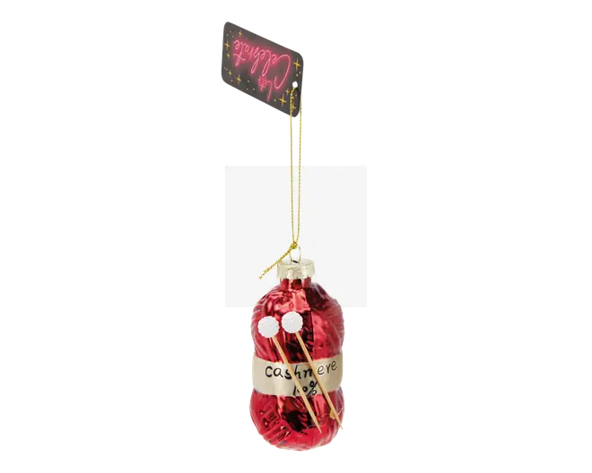 En blank rød julekugle formet som et garnnøgle, omviklet med en beige etiket, hvor der står "cashmere 100%" med håndskrevet tekst. To små strikkepinde med hvide pomponer i toppen er stukket ind i garnet, hvilket understreger strikketemaet. Ornamentet har en gylden ophængssnor og et sort gavemærke med festlig skrift fastgjort øverst.