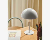 En moderne bordlampe med en blank, hvid, kuppelformet skærm og en slank, lige stander på en rund fod. Lampen har et minimalistisk design med glatte overflader og rene linjer. Den står på et lyst træskrivebord ved siden af en kop med farveblyanter og en skitsebog med en tegning. Lampens ledning er synlig og løber fra foden.