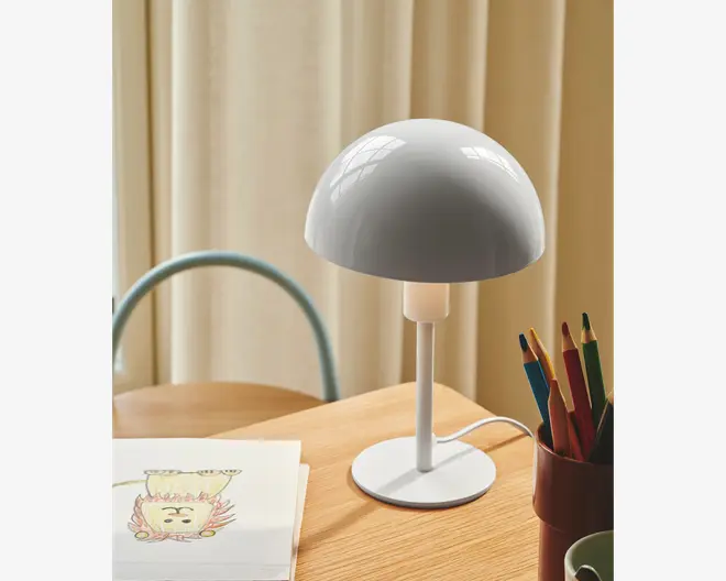 En moderne bordlampe med en blank, hvid, kuppelformet skærm og en slank, lige stander på en rund fod. Lampen har et minimalistisk design med glatte overflader og rene linjer. Den står på et lyst træskrivebord ved siden af en kop med farveblyanter og en skitsebog med en tegning. Lampens ledning er synlig og løber fra foden.