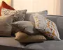 En gruppe dekorative puder i blandede ტექsturer og mønstre på en grå sofa, herunder en rustfarvet velourpude, blomstermønster, beige striber, en ensfarvet taupe pude, en tekstureret tan pude og en hvid pude med et orange geometrisk mønster.