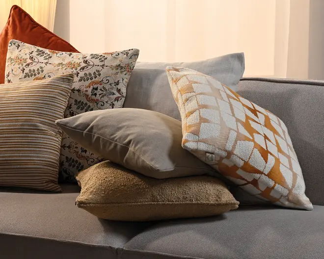 En gruppe dekorative puder i blandede ტექsturer og mønstre på en grå sofa, herunder en rustfarvet velourpude, blomstermønster, beige striber, en ensfarvet taupe pude, en tekstureret tan pude og en hvid pude med et orange geometrisk mønster.