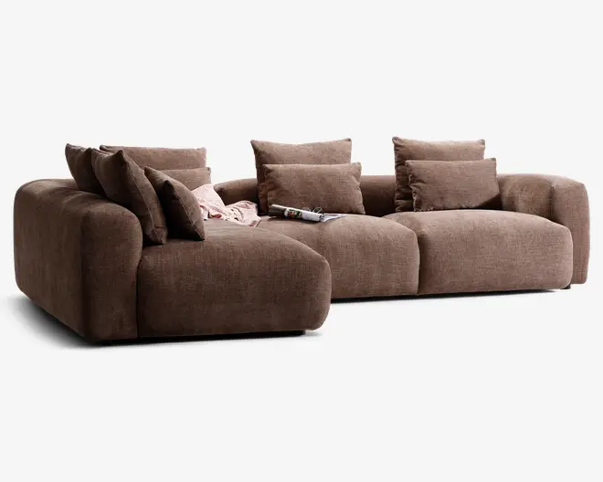 En moderne, modulopbygget sofa med dybt sæde og brede armlæn, betrukket med et tekstureret brunt stof. Sofaen har bløde, fyldige ryg- og sædehynder for ekstra komfort og leveres med flere matchende pyntepuder. Der ligger et lyst tæppe og et magasin afslappet på sædet, hvilket giver et hyggeligt og indbydende udtryk.