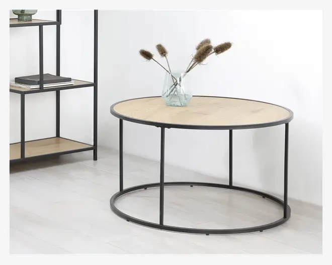 Rundt bord med en lys træbordplade og sort metalstel. Bordet har et minimalistisk design med fire slanke ben, der er forbundet af en cirkulær bund. På bordet står en klar glasvase med tørrede græsser. Bordet står på et lyst trægulv, og en matchende reol ses i baggrunden.