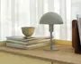 En lille, moderne bordlampe med mat lysegrå finish. Lampen har en kuppelformet skærm og en slank cylindrisk stang, der er fastgjort til en rund fod. Den står på en træoverflade ved siden af en stak magasiner og en lille beige skål, med blødt naturligt lys, der filtrerer gennem gennemsigtige gardiner i baggrunden.