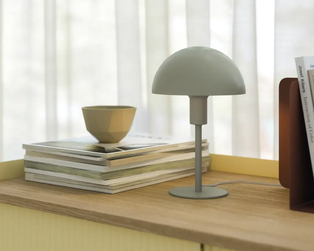 En lille, moderne bordlampe med mat lysegrå finish. Lampen har en kuppelformet skærm og en slank cylindrisk stang, der er fastgjort til en rund fod. Den står på en træoverflade ved siden af en stak magasiner og en lille beige skål, med blødt naturligt lys, der filtrerer gennem gennemsigtige gardiner i baggrunden.