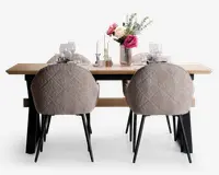 Et moderne spisebordssæt med et rektangulært bord i lyst træ og sorte metalben. Fire polstrede stole med tekstureret, lysebeige stof og sorte metalben omkranser bordet. Bordet er stilfuldt dækket med hvide tallerkener, vinglas og sølvbestik, og er pyntet med en blomsterdekoration i en rillet glasvase samt høje hvide lys i stager.