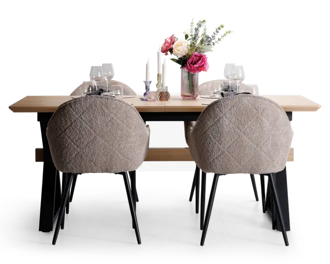 Et moderne spisebordssæt med et rektangulært bord i lyst træ og sorte metalben. Fire polstrede stole med tekstureret, lysebeige stof og sorte metalben omkranser bordet. Bordet er stilfuldt dækket med hvide tallerkener, vinglas og sølvbestik, og er pyntet med en blomsterdekoration i en rillet glasvase samt høje hvide lys i stager.