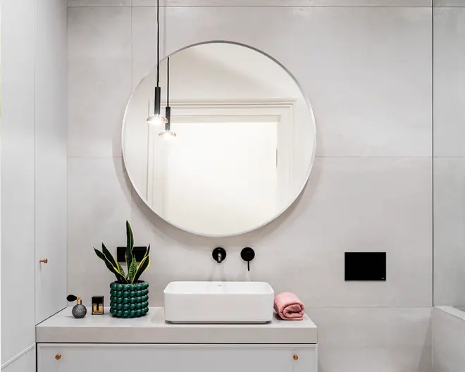 Et moderne badeværelsesbord med en stor rund spejl monteret på en lysegrå flisevæg. Bordpladen er hvid med en rektangulær fritstående vask og mat sort vægmonteret armatur. Til venstre på bordet står en dekorativ grøn vase med en slangeplante, et lille stearinlys og en grå ornament med tekstur. Til højre ligger et pænt sammenfoldet lyserødt håndklæde. To minimalistiske pendellamper med synlige pærer hænger fra loftet over vasken og giver et stilfuldt udtryk.