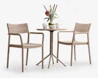 Sættet består af to beige stole med armlæn og et rundt bord med en slank, firebenet fod. Stolene har et moderne, minimalistisk design med let buede rygge og sæder. Bordet er pyntet med en vase med blomster, to krus og en skål med grønne æbler, hvilket giver et hyggeligt indtryk til afslappet spisning eller kaffe.