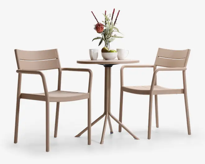 Sættet består af to beige stole med armlæn og et rundt bord med en slank, firebenet fod. Stolene har et moderne, minimalistisk design med let buede rygge og sæder. Bordet er pyntet med en vase med blomster, to krus og en skål med grønne æbler, hvilket giver et hyggeligt indtryk til afslappet spisning eller kaffe.