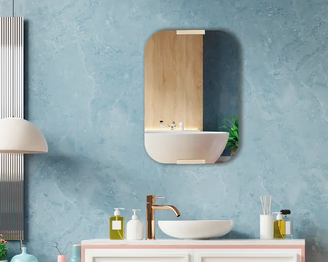 Moderne badeværelsesindretning med en rund, hvid håndvask ovenpå bordet og en slank, høj vandhane i rosaguld. På bordpladen står en sæbedispenser, en lotionflaske, en duftspreder med pinde og en lille flaske flydende sæbe, alle i minimalistisk design. Bagvæggen har en lyseblå, tekstureret overflade, og over vasken hænger et rektangulært spejl med afrundede hjørner og metalbeslag foroven og forneden. Spejlet reflekterer en træpanelvæg og en grøn potteplante, hvilket tilfører et naturligt og varmt præg til det moderne udtryk.