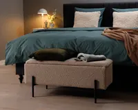Rektangulær bænk med blødt, tekstureret beige betræk og sorte metalben. Bænken står for enden af en seng og har et hyggeligt, indbydende udseende. Ovenpå bænken ligger en mørkegrøn velourpude og et sammenfoldet grå- og hvidstribet tæppe. Bænkens design er moderne og minimalistisk og passer godt til det nutidige soveværelsesindretning.
