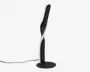 Moderne lampe med et snoet, skulpturelt design. Lampen har en mat sort overflade og en sammenhængende hvid LED-lysbånd, der snor sig rundt om kroppen. Den står på en rund, flad fod og har en slank, lodret profil, hvilket gør den velegnet til moderne indretning.