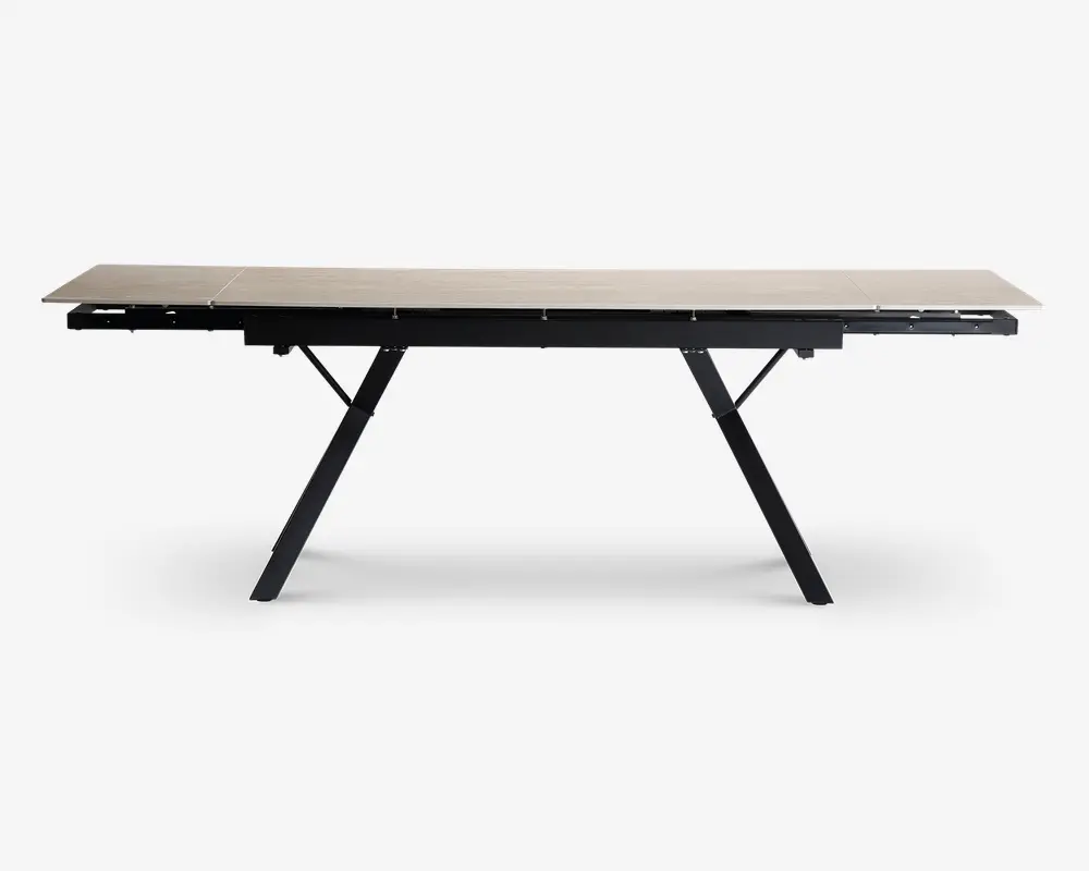 Rektangulært bord med en lys, glat bordplade og sorte, skråtstillede metalben. Bordet har et moderne, minimalistisk design og ser ud til at være udtrækkeligt, hvilket ses på de synlige samlinger og udtræksmekanismen under bordpladen.