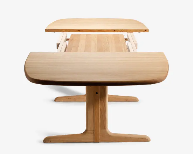 Bord i lyst træ med centralt søjleben, vist i udtrukket position hvor bordpladen er delt i to sektioner med plads i midten til en ekstra plade. Bordpladens kanter er afrundede, og træets årer er synlige, hvilket giver et naturligt udtryk.