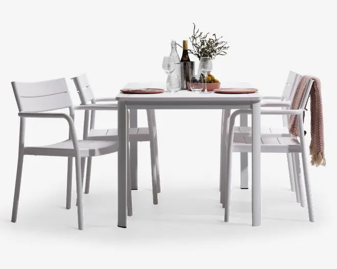 Et moderne spisesæt med et rektangulært bord og fire matchende stole, alle i en elegant hvid finish. Stolene har armlæn og lamelrygge for komfort og stil. Bordet er dækket med lyserøde dækkeservietter, glas og en midterdekoration bestående af en vase med blomster, flasker og en skål med frugt. En af stolene har et sammenfoldet tæppe hængende over ryglænet, hvilket giver et hyggeligt udtryk.