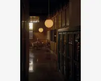 Et svagt oplyst, elegant spiseområde med vægge af træpaneler og blanke gulve. Rummet har runde, matterede glaspendellamper, der hænger fra loftet og udsender et varmt lys. I baggrunden ses et dækket spisebord med stole, stearinlys og service, hvilket skaber en indbydende og intim stemning. Store vitrineskabe med glaslåger står langs den ene side af gangen.