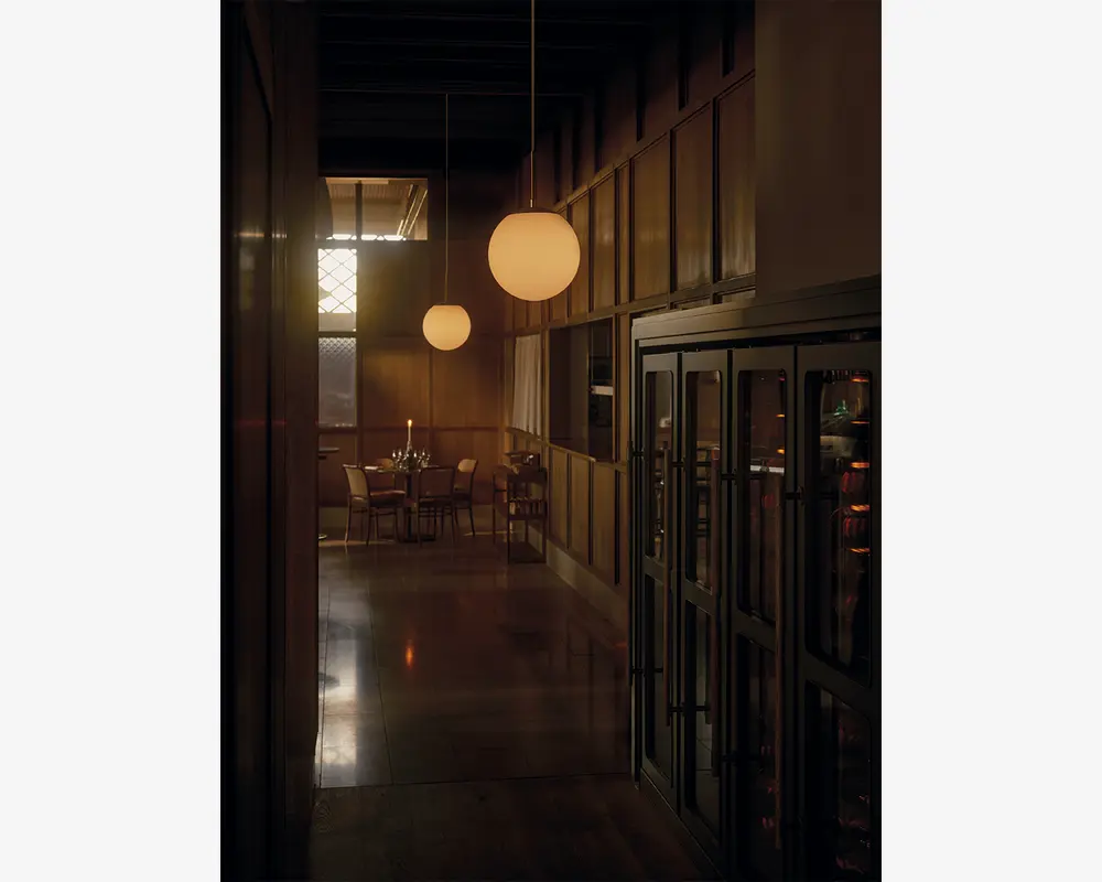 Et svagt oplyst, elegant spiseområde med vægge af træpaneler og blanke gulve. Rummet har runde, matterede glaspendellamper, der hænger fra loftet og udsender et varmt lys. I baggrunden ses et dækket spisebord med stole, stearinlys og service, hvilket skaber en indbydende og intim stemning. Store vitrineskabe med glaslåger står langs den ene side af gangen.