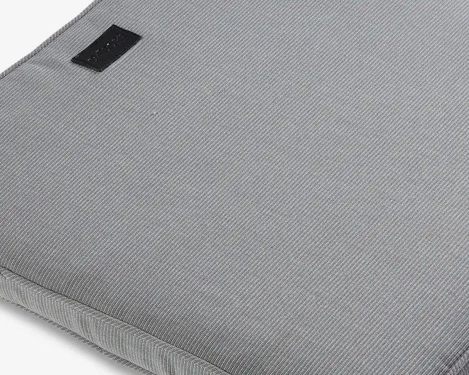 Nærbillede af en lys grå stofoverflade med et diskret diagonalt vævemønster. Kanterne er pænt syet, og der er et lille sort rektangulært mærke syet fast nær det ene hjørne. Materialet virker slidstærkt og let polstret, hvilket antyder, at det giver en vis beskyttelse af indholdet.