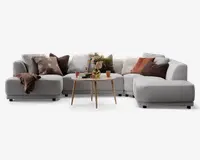 En moderne, L-formet sofa med lysegråt betræk og afrundede kanter. Sofaen er pyntet med forskellige pyntepuder i brune, rustfarvede og olivengrønne nuancer, som har forskellige teksturer og mønstre. Et blødt, brunt tæppe er lagt over det ene armlæn. Foran sofaen står et rundt sofabord i lyst træ med en dekorativ vase med blomster, to krus og et lille fad, hvilket skaber en hyggelig og indbydende stemning.