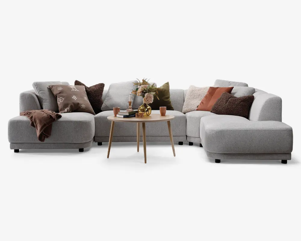 En moderne, L-formet sofa med lysegråt betræk og afrundede kanter. Sofaen er pyntet med forskellige pyntepuder i brune, rustfarvede og olivengrønne nuancer, som har forskellige teksturer og mønstre. Et blødt, brunt tæppe er lagt over det ene armlæn. Foran sofaen står et rundt sofabord i lyst træ med en dekorativ vase med blomster, to krus og et lille fad, hvilket skaber en hyggelig og indbydende stemning.