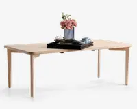 Rektangulært bord med lys træfinish, afrundede hjørner og slanke, tilspidsede ben. Bordpladen er dekoreret med en rillet vase med lyserøde blomster, et sort bakke, en sammenfoldet serviet, en kop med underkop og en lille skål, hvilket giver et moderne og minimalistisk udtryk.