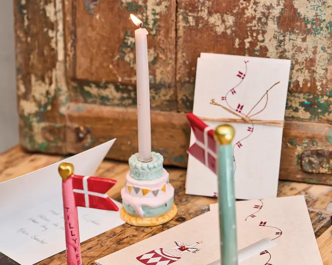 Billedet viser en dekorativ opstilling med en lysholder formet som en lagkage i pastelfarver, hvor der står et tændt hvidt stearinlys. Der er to små flagstænger med røde og hvide flag, hver med et gyldent hjerte på toppen, og på det ene flag står der 'TILLYKKE'. Flere lykønskningskort er lagt frem, nogle med illustrationer af flag og festlige motiver, og et kort er bundet med en enkel snor. Baggrunden består af en rustik, slidt træoverflade og et gammelt skab, hvilket giver opstillingen et vintagepræg.