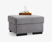 Firkantet puf betrukket med lysegråt tekstureret stof, med et rent og moderne design med lige kanter og sorte blokfødder. Den øverste overflade er flad og robust, velegnet som bord, hvilket vises med en træbakke med en vandflaske, glas og frugt. Puffen ser ud til at have et rummeligt indre, muligvis til opbevaring.