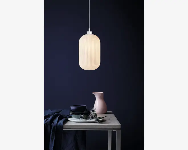 En pendellampe med en lodret rillet, mat hvid glasskærm i en aflang oval form. Lampen udsender et blødt, varmt lys og hænger i en tynd hvid ledning. Designet er minimalistisk og moderne, velegnet til nutidige indretninger.
