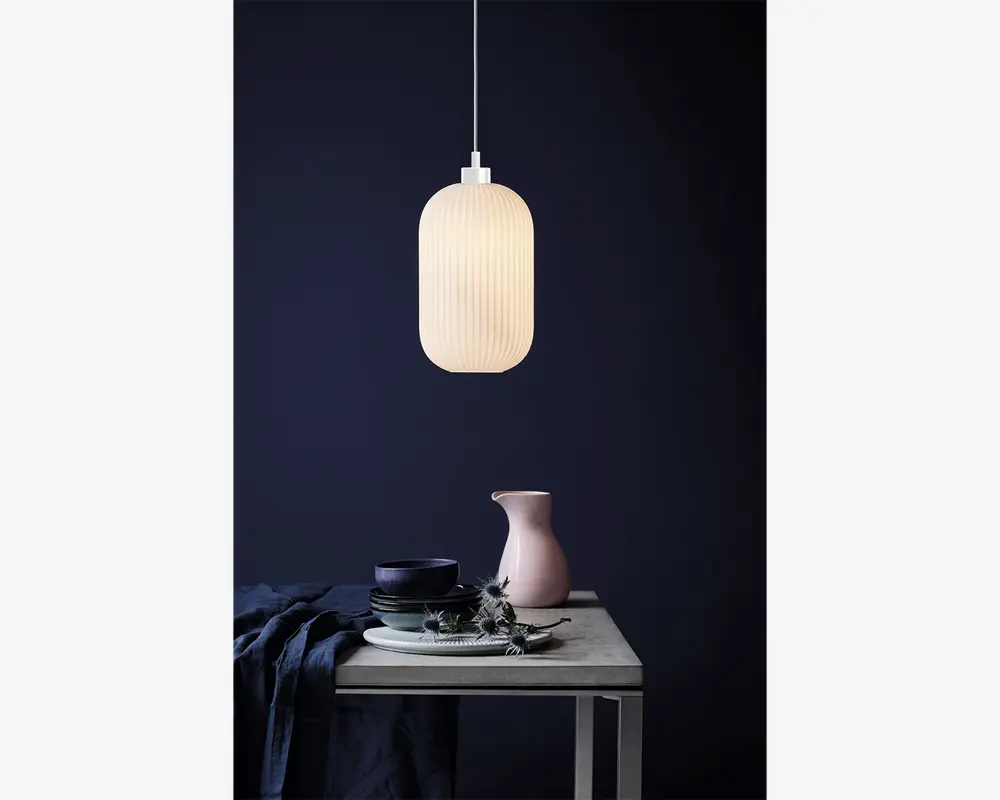 En pendellampe med en lodret rillet, mat hvid glasskærm i en aflang oval form. Lampen udsender et blødt, varmt lys og hænger i en tynd hvid ledning. Designet er minimalistisk og moderne, velegnet til nutidige indretninger.