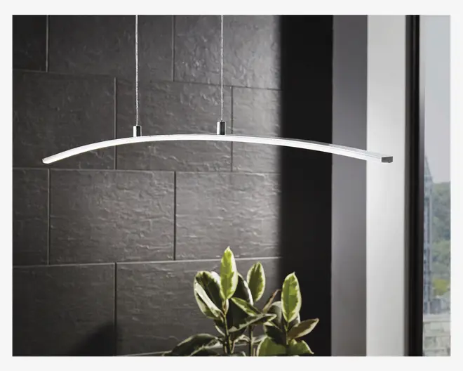 Et moderne pendellampe-armatur med et slankt, let buet stangdesign i metalfinish. Lampen hænger ned fra loftet i to tynde kabler, hvilket giver et minimalistisk og nutidigt udtryk. Lyskilden løber langs undersiden af den buede stang og giver jævn belysning. Baggrunden viser en mørk, tekstureret væg og et vindue, med en grøn plante synlig under lampen.