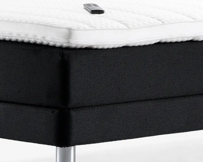 Nærbillede af et sengehjørne med en tyk, struktureret hvid topmadras med synlig lynlås, placeret på en solid sort polstret sengebund. Bunden har en vævet stofstruktur og understøttes af et blankt metalben. En lille fjernbetjening ligger ovenpå topmadrassen.