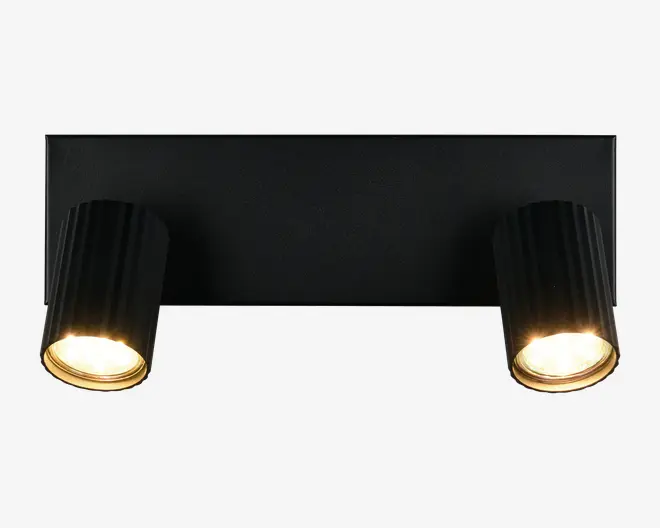 Et moderne vægmonteret armatur med to justerbare cylindriske spotlys, der er fastgjort til en rektangulær base. Spotlysene har en mat sort finish med diskrete lodrette riller for tekstur, og de indvendige kanter er guldfarvede, hvilket fremhæver det varme lys. Armaturets minimalistiske design gør det velegnet til moderne indretning, og de justerbare hoveder giver mulighed for retningsbestemt belysning.