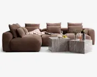 Et moderne stuearrangement med en stor, blød brun hjørnesofa med dybe sæder og flere store ryg- og sidepuder for ekstra komfort. Sofaen har et blødt, tekstureret stof og er pyntet med et lyserødt tæppe. Foran sofaen står to firkantede sofaborde i grå marmor med naturligt stenlook. Bordene er dekoreret med et vinglas, en vinflaske, en skål snacks, en tallerken med frugt og en klar vase med blomster, hvilket skaber en hyggelig og indbydende stemning.