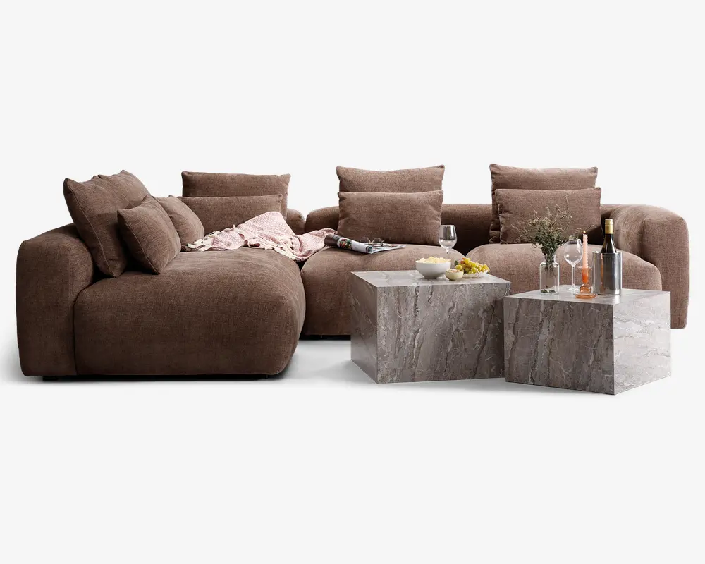 Et moderne stuearrangement med en stor, blød brun hjørnesofa med dybe sæder og flere store ryg- og sidepuder for ekstra komfort. Sofaen har et blødt, tekstureret stof og er pyntet med et lyserødt tæppe. Foran sofaen står to firkantede sofaborde i grå marmor med naturligt stenlook. Bordene er dekoreret med et vinglas, en vinflaske, en skål snacks, en tallerken med frugt og en klar vase med blomster, hvilket skaber en hyggelig og indbydende stemning.