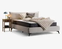 Polstret seng med høj, blød sengegavl i lysebeige stof. Sengen har en tyk, mørkegrå madras og en hvid topmadras. Sengetøjet består af en beige og hvidstribet dyne, flere pyntepuder i forskellige former og neutrale farver samt et matchende pudebetræk. Ved siden af sengen står et natbord i træ med skuffe og hylde, hvorpå der står en blå lampe, en lille vase med blomster og en kop.